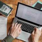High-angle view of hands typing on a laptop surrounded by books and papers., tags: art persuasive - pexel