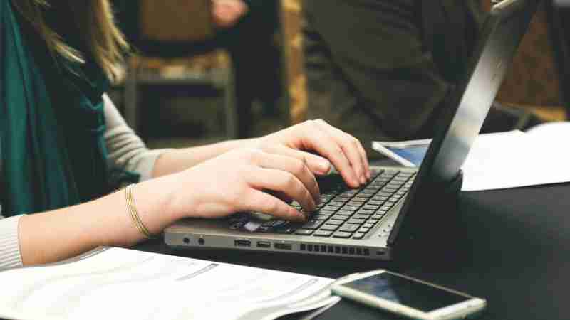 Adult woman typing on a laptop at a desk with a smartphone and documents., tags: digital - pexel