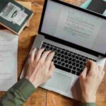 High-angle view of hands typing on a laptop surrounded by books and papers., tags: persuasive copy modern - pexel