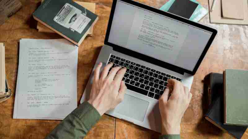 High-angle view of hands typing on a laptop surrounded by books and papers., tags: persuasive copy modern - pexel